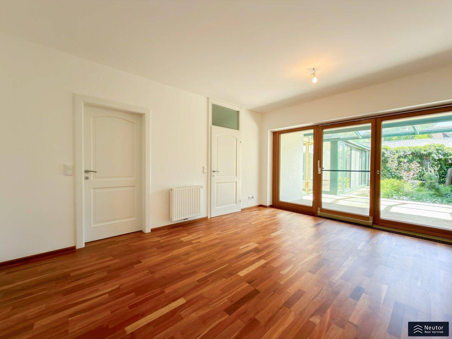 Geräumiges Zimmer mit Holzboden und großen Glasfronten zum Garten, sehr hell.
