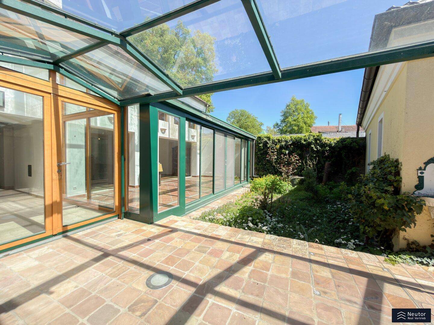 Sonniger Terrassenbereich mit Terrakottafliesen und Glasüberdachung, umgeben von Grün.