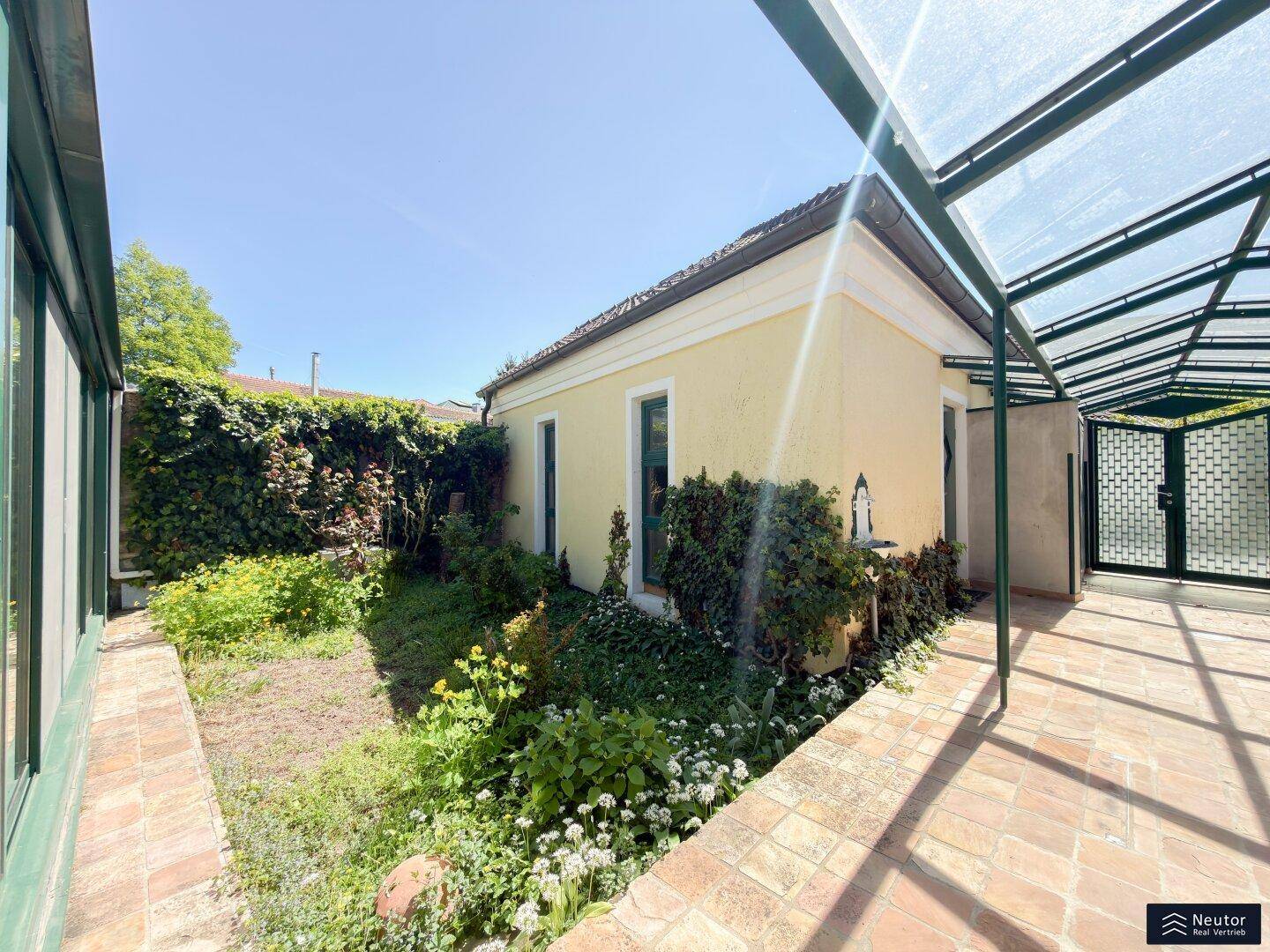 Sonniger Gartenweg entlang des Hauses mit üppiger Bepflanzung und Terrakottafliesen.