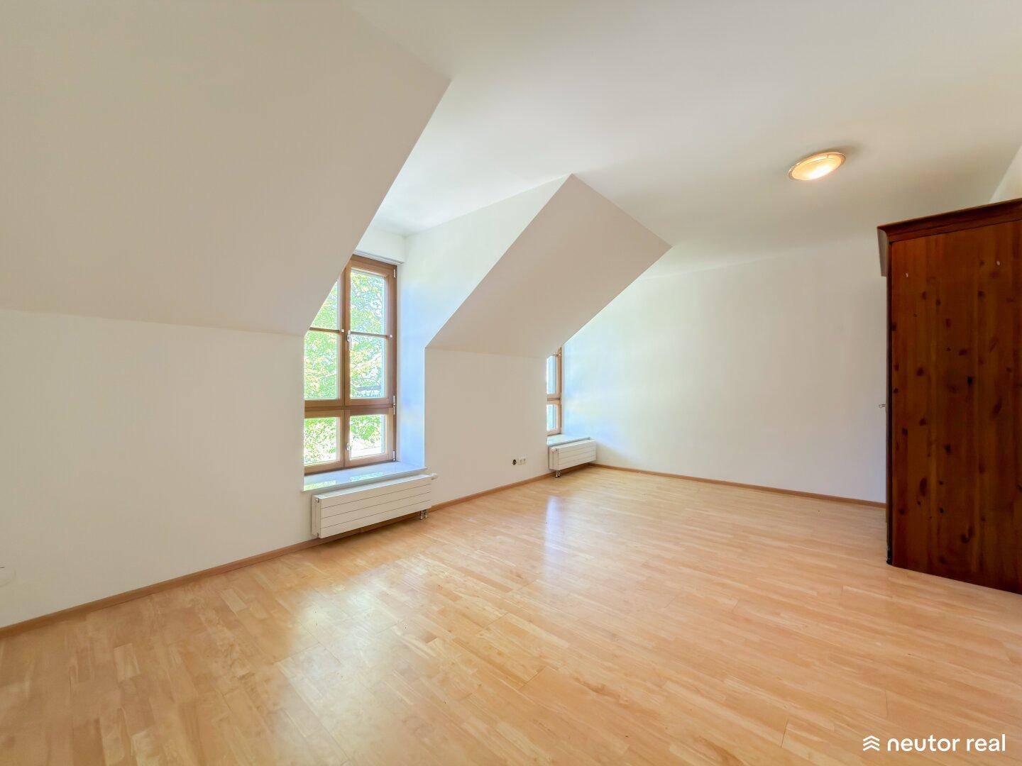 Helles Zimmer im Dachgeschoss mit Holzboden, schrägen Wänden und großen Fenstern.