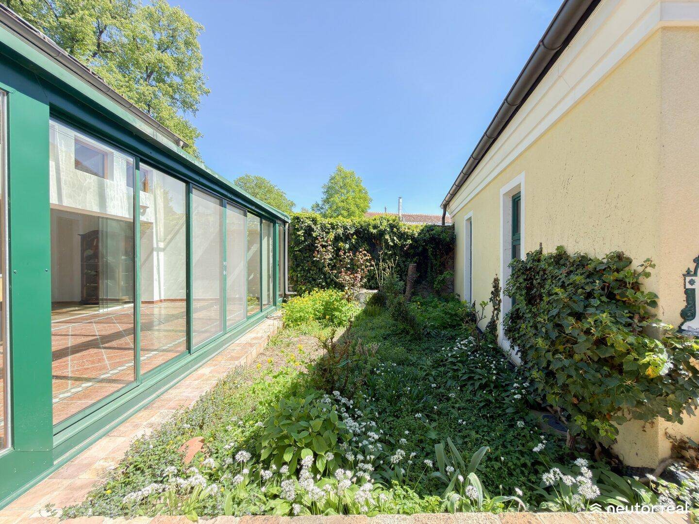 Schmaler Gartenweg zwischen dem Haus und einem verglasten Anbau mit üppigem Grün und viel Sonne.