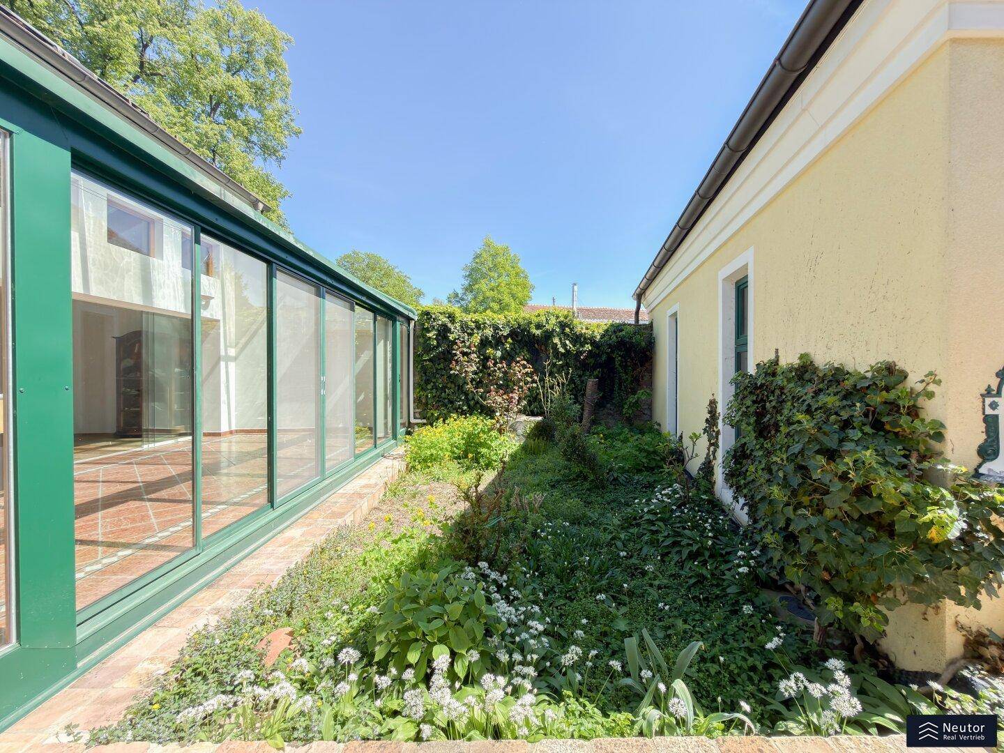 Schmaler Gartenbereich zwischen Wintergarten und Hauswand mit üppiger Bepflanzung.