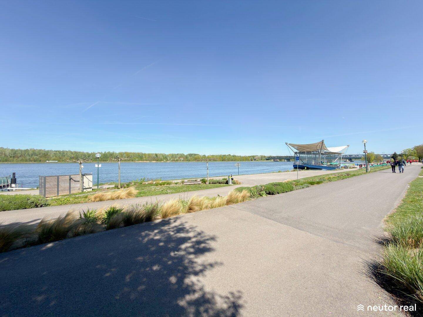 Weitläufiger Blick auf einen Fluss mit Uferpromenade und Spazierweg an einem sonnigen Tag.