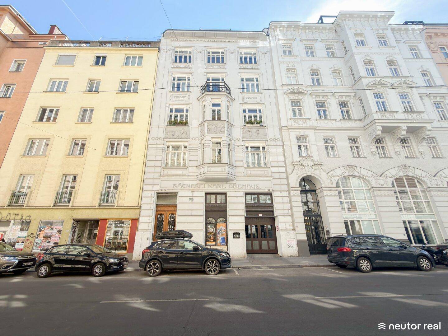 Historische Fassade eines Mehrfamilienhauses mit Geschäften im Erdgeschoss und parkenden Autos davor.