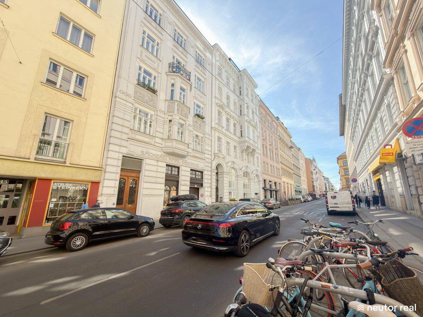 Belebtes Stadtbild mit historischen Gebäuden, parkenden Autos und Fahrrädern an einem sonnigen Tag.