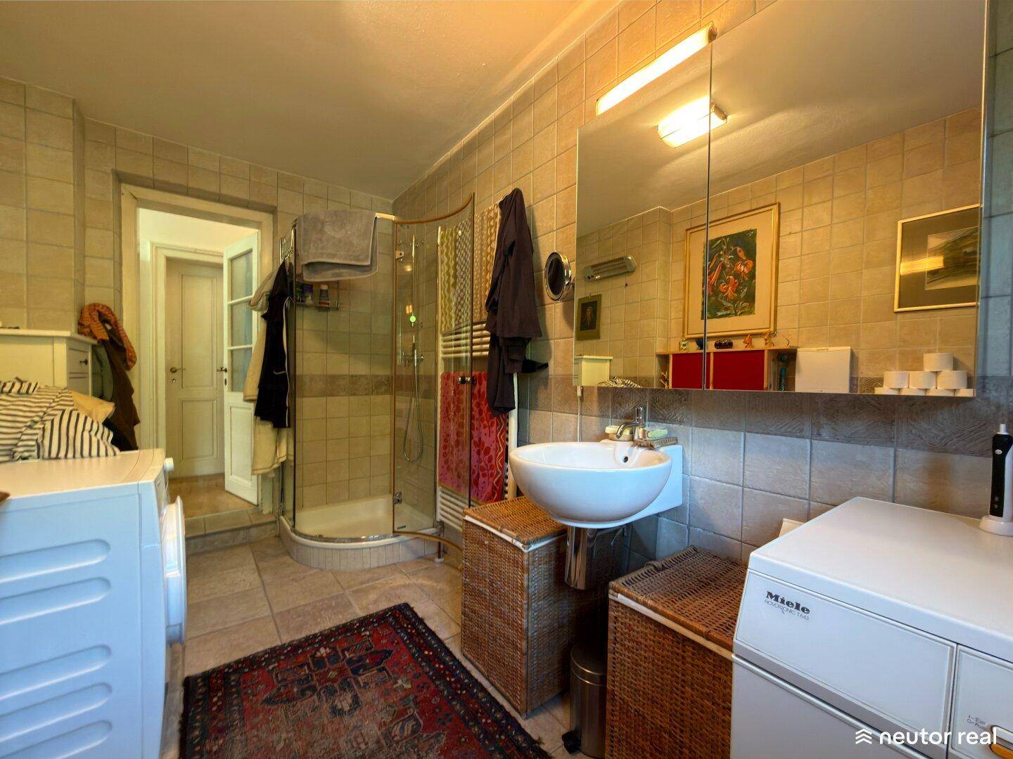 Gefliestes Badezimmer mit Dusche, Waschmaschine und Spiegel über dem Waschbecken.