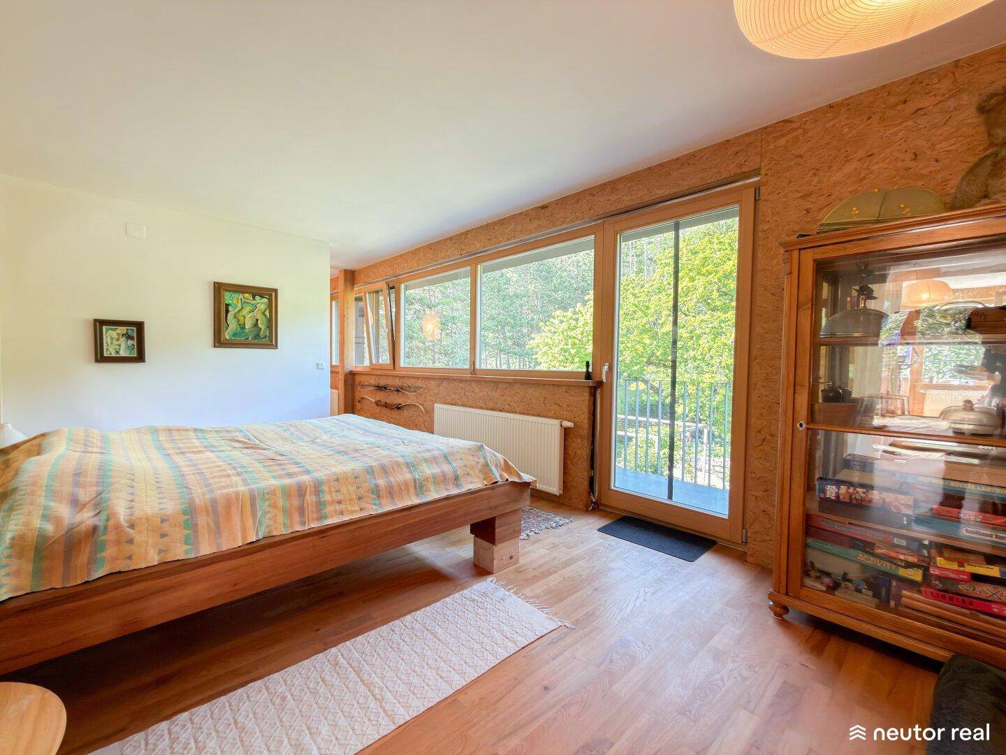 Schlafzimmer mit Holzbett, Holzboden und großen Fenstern mit Blick auf die Natur.