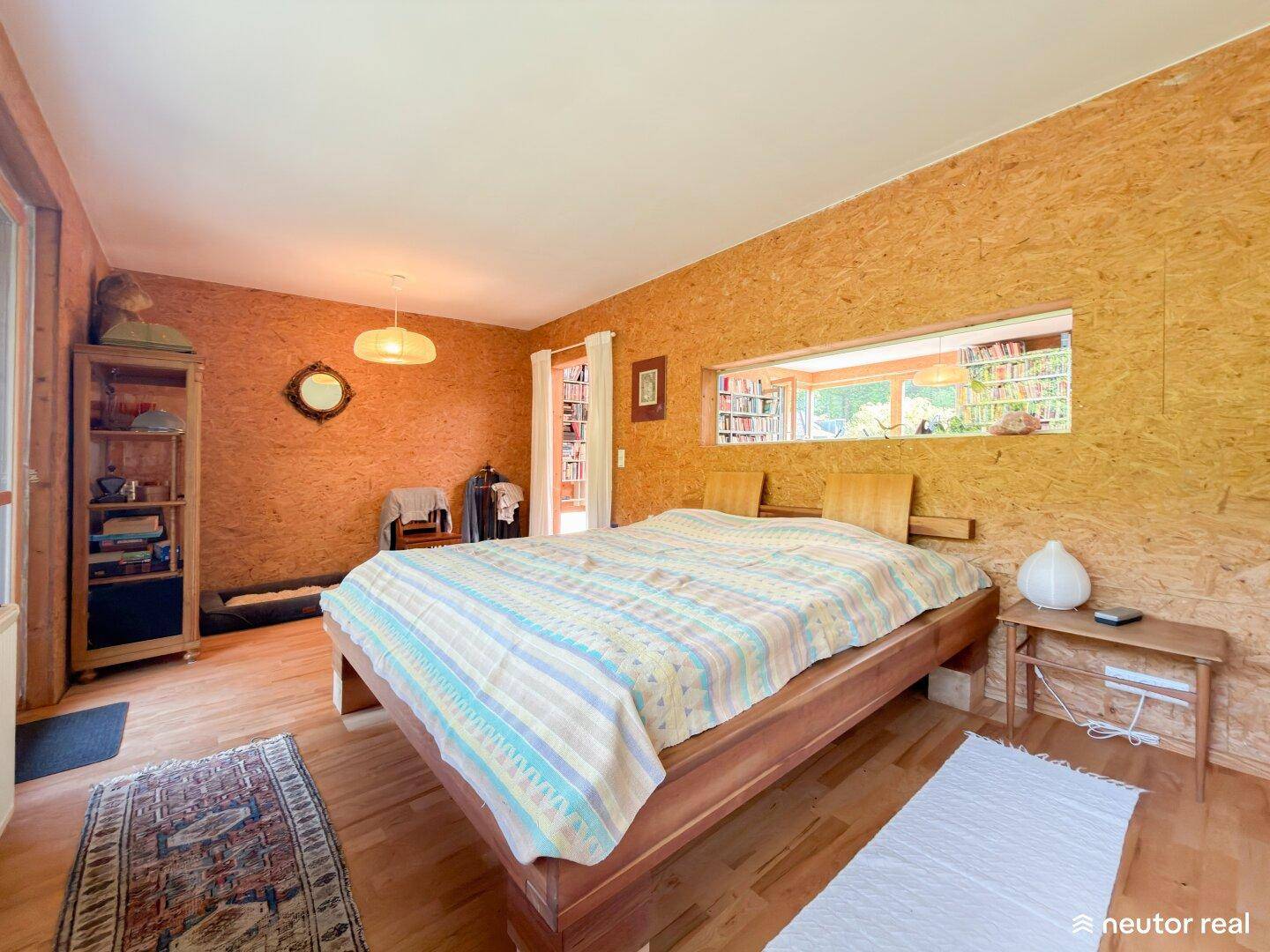 Schlafzimmer mit Holzbett, Holzboden und einem Fenster, das den Blick in einen angrenzenden Raum freigibt.