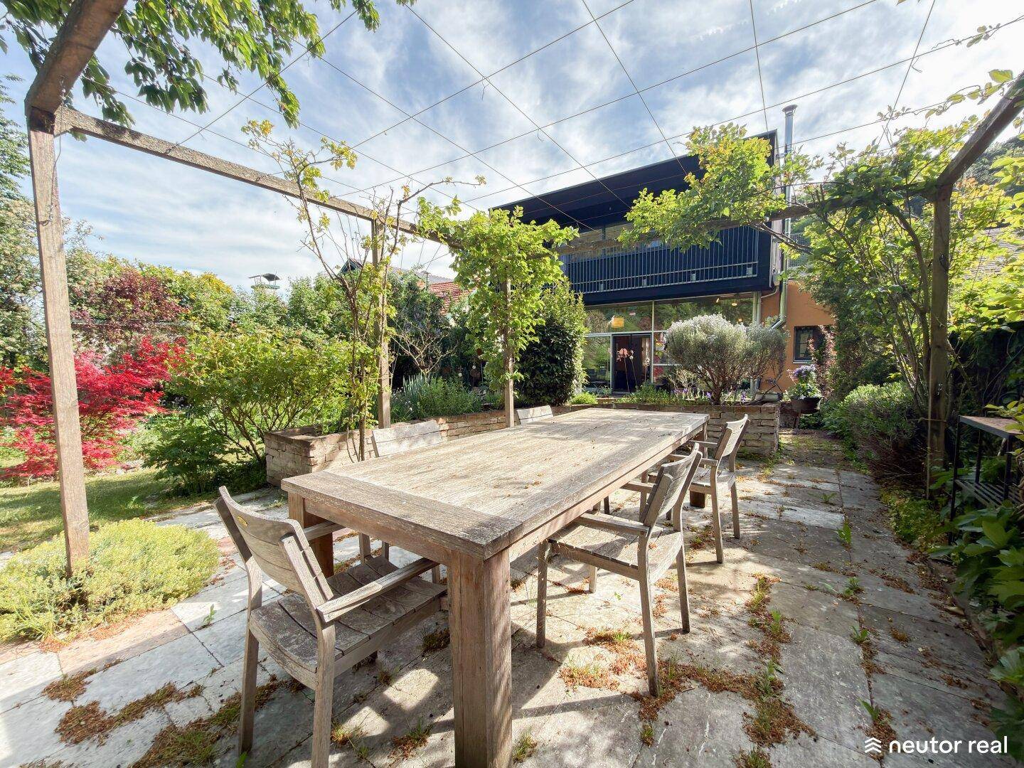 Gemütlicher Essbereich im Freien auf einer gepflasterten Terrasse mit Holztisch und Blick auf das moderne Haus.