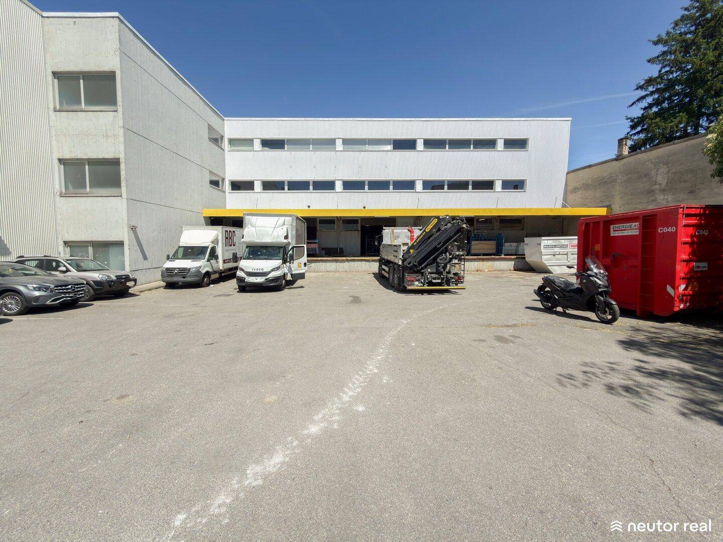 Parkplatz und Ladezone vor einem Gewerbegebäude mit gelben Akzenten an einem sonnigen Tag.