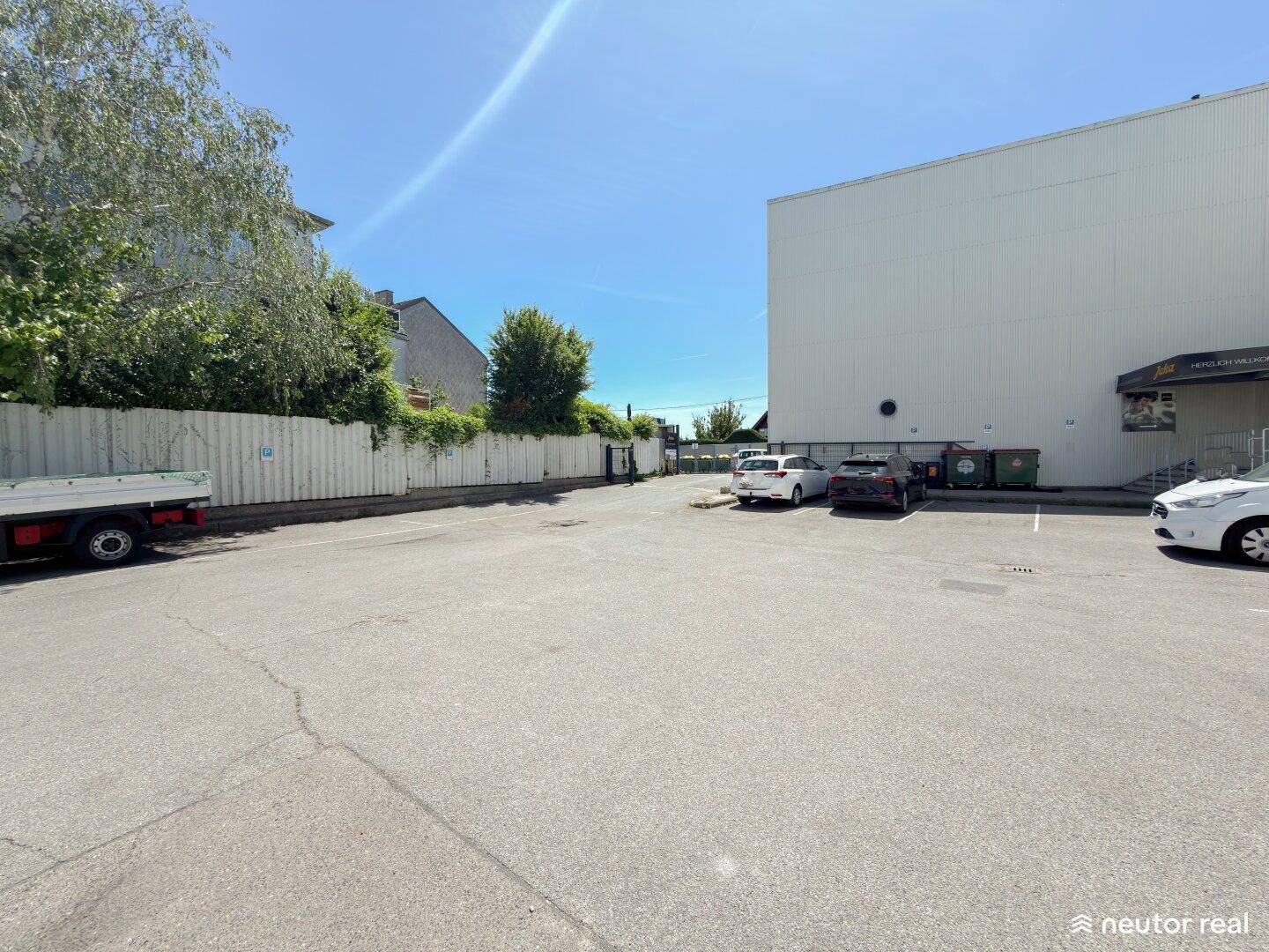 Großer, asphaltierter Parkplatz vor einem Industriegebäude mit einem Zaun und Bäumen im Hintergrund.