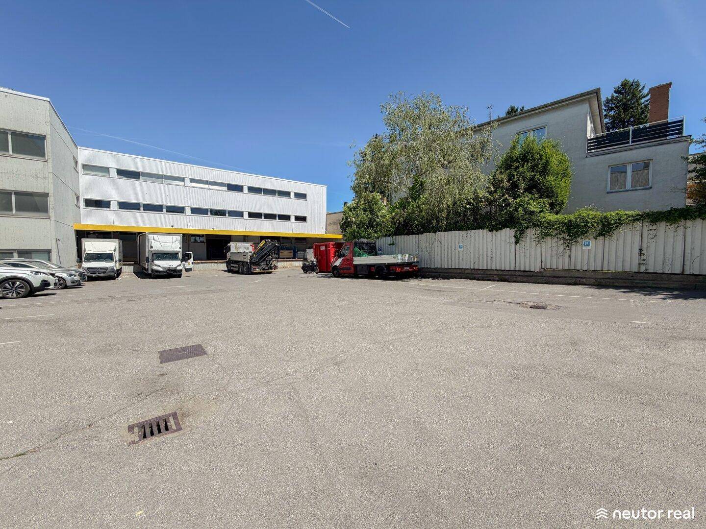 Parkplatzbereich vor dem Gewerbegebäude mit Lieferfahrzeugen und Bäumen im Hintergrund.