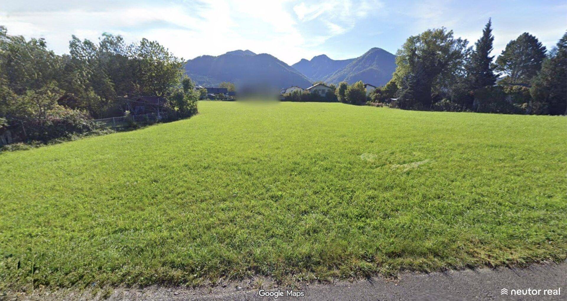 Unverbaute grüne Fläche umgeben von Bäumen und einer beeindruckenden Bergkulisse im Hintergrund.