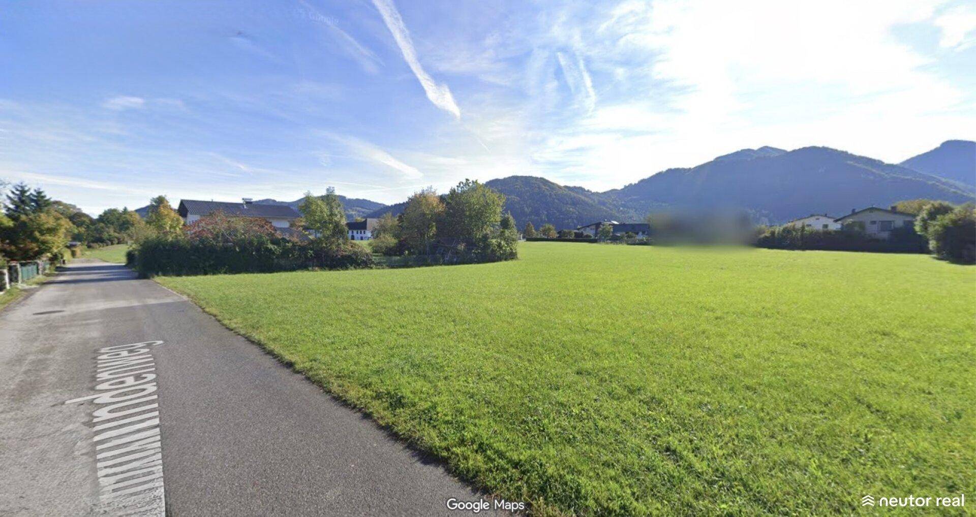 Weitläufige grüne Wiese entlang einer Straße mit Häusern und Bergen in der Ferne.