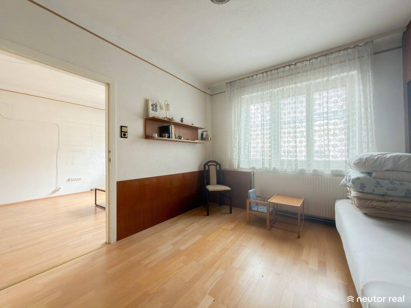 Schlafzimmer mit hellem Holzboden, großem Fenster und einfacher Einrichtung.