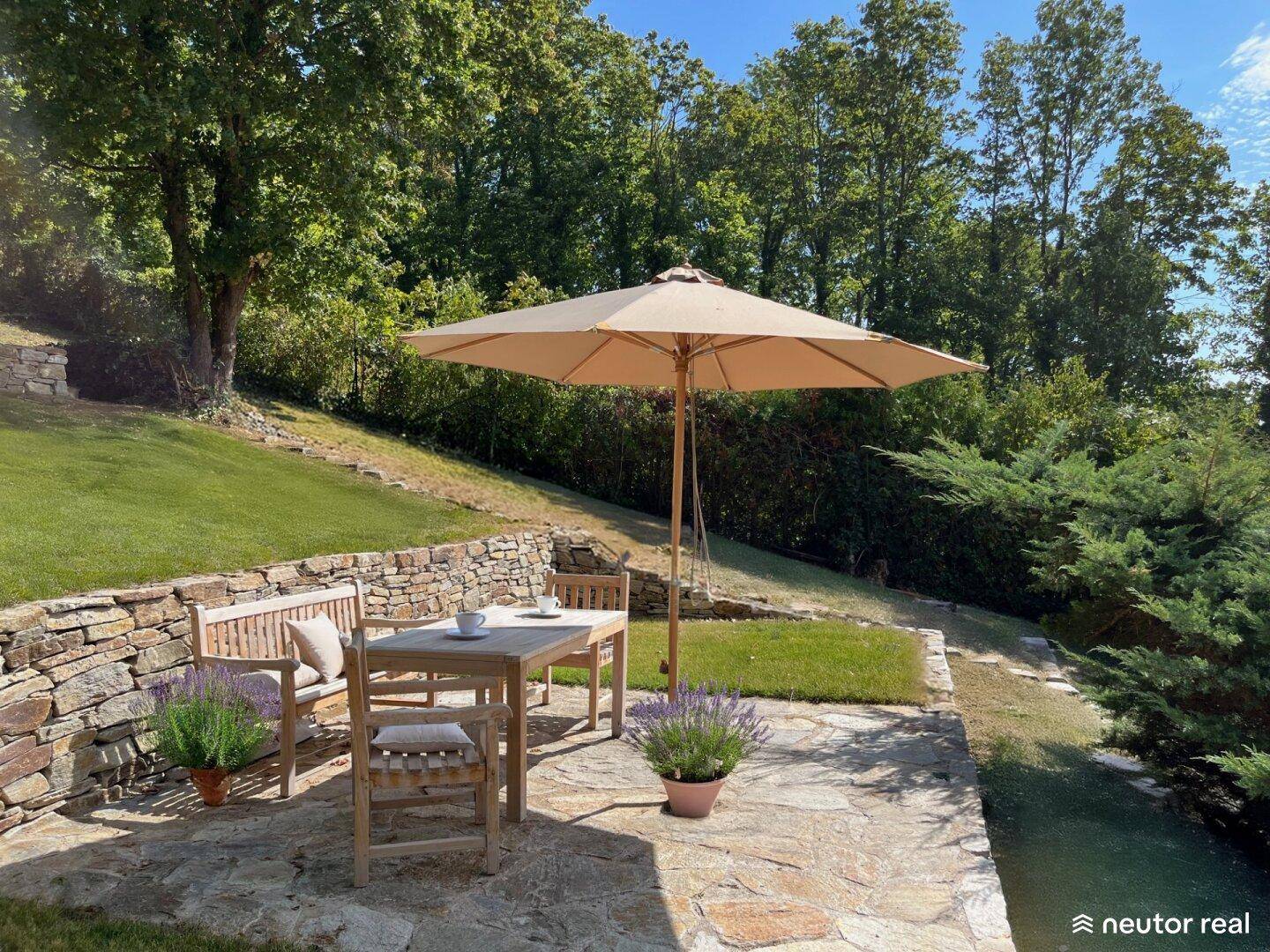 Sonniger Terrassenbereich mit Steinfliesen, Holztisch und Blick auf den grünen Garten.