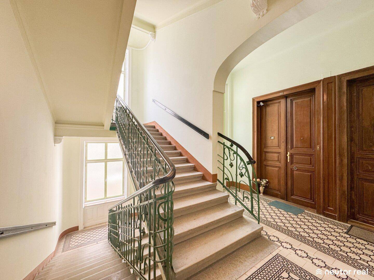 Historisches Treppenhaus mit kunstvollem Geländer, hohen Decken und gemustertem Boden.