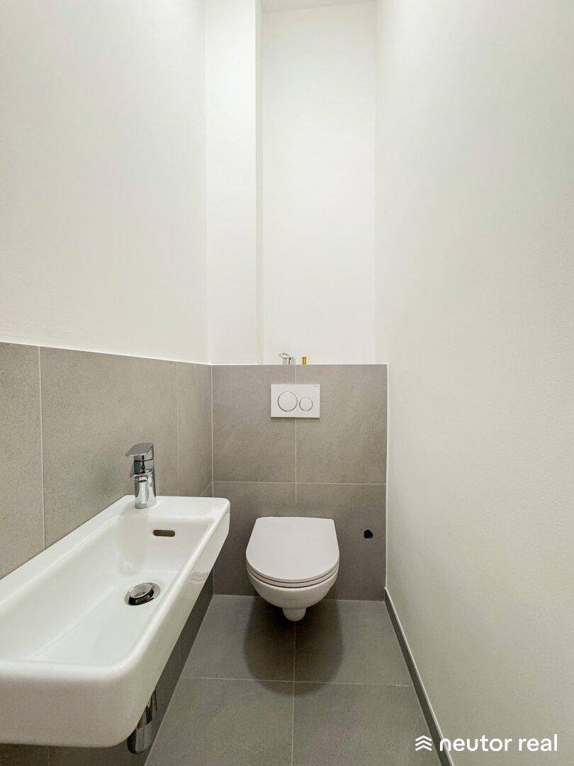 Modernes Gäste-WC mit Wand-WC und schmalem Waschbecken, in hellen Grautönen gehalten.