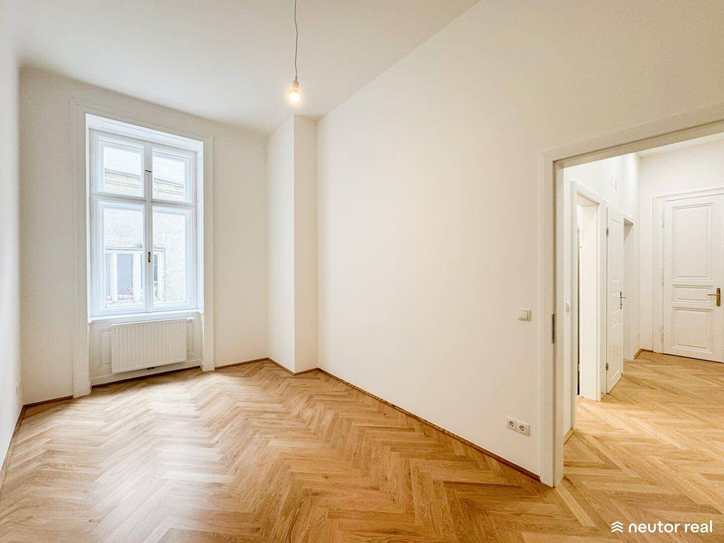 Helles, leeres Zimmer mit Fischgrätparkett, großem Fenster und Zugang zu weiteren Räumen.
