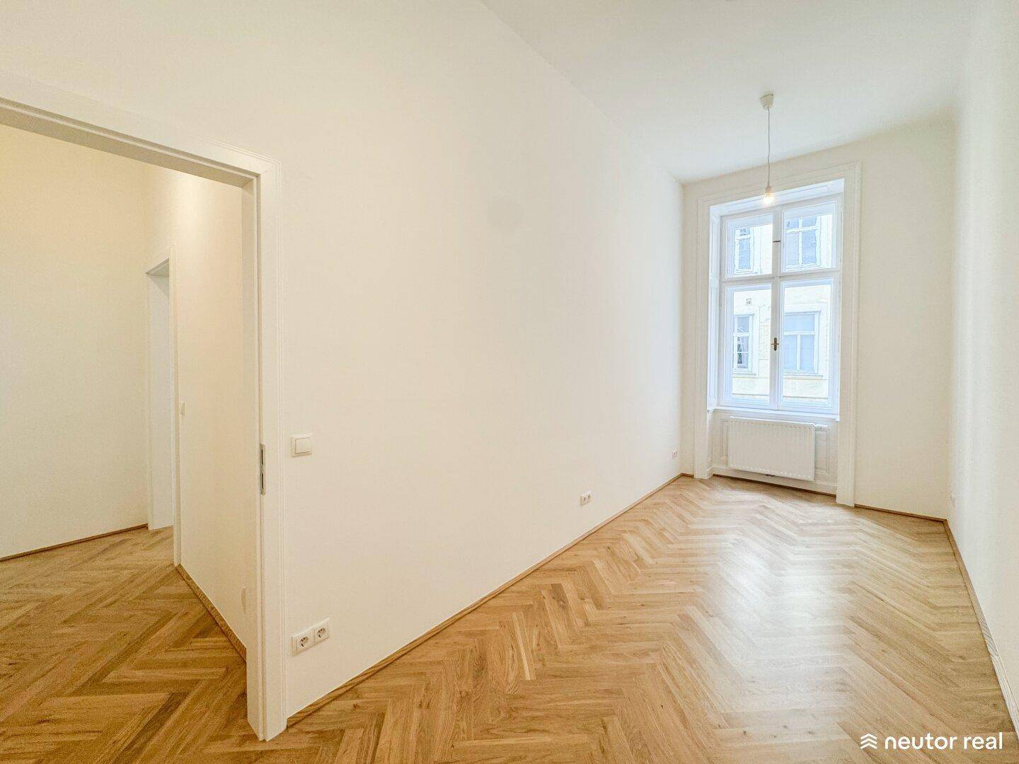 Helles, leeres Zimmer mit Fischgrätparkett, großem Fenster und Zugang zu weiteren Bereichen.