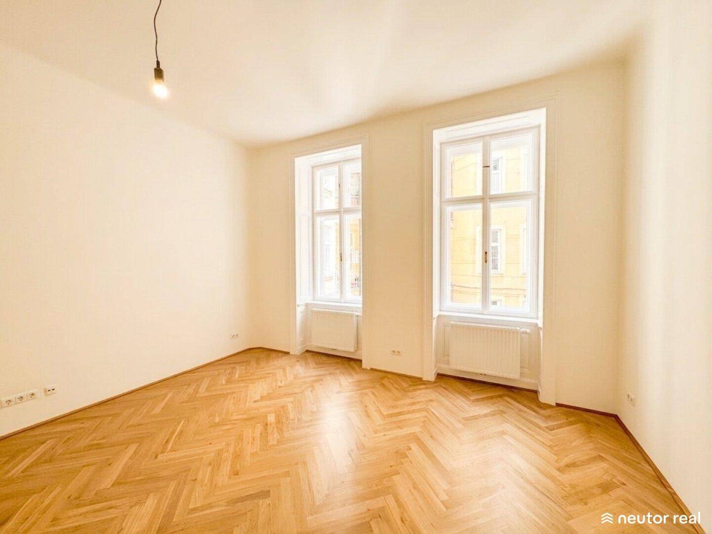 Helles, leeres Zimmer mit zwei großen Fenstern, Fischgrätparkett und viel Tageslicht.