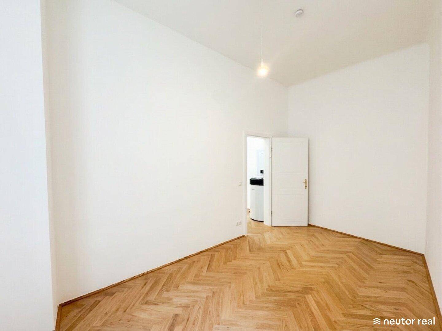 Leeres Zimmer mit Fischgrätparkett und Zugang zu einem Bereich mit Heizungsanlage.