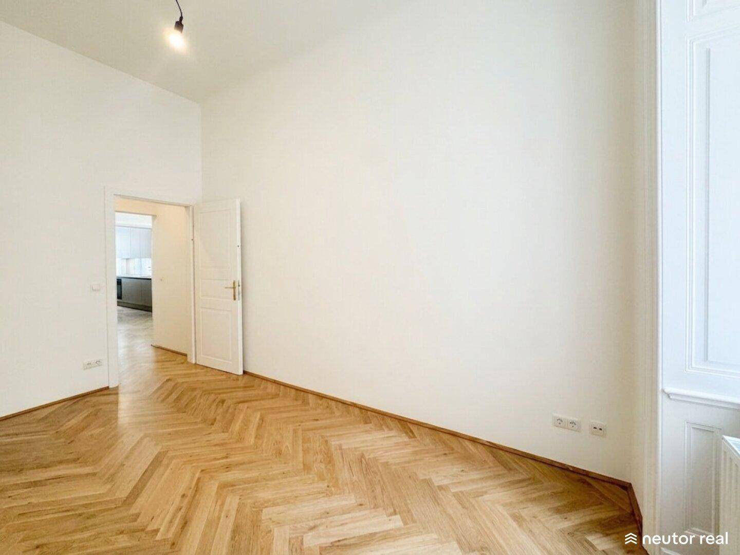 Geräumiges, leeres Zimmer mit Fischgrätparkett und Zugang zur modernen Küche.