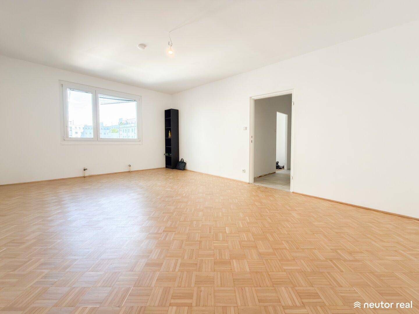 Großes, leeres Wohnzimmer mit Fischgrätparkett und viel Tageslicht.
