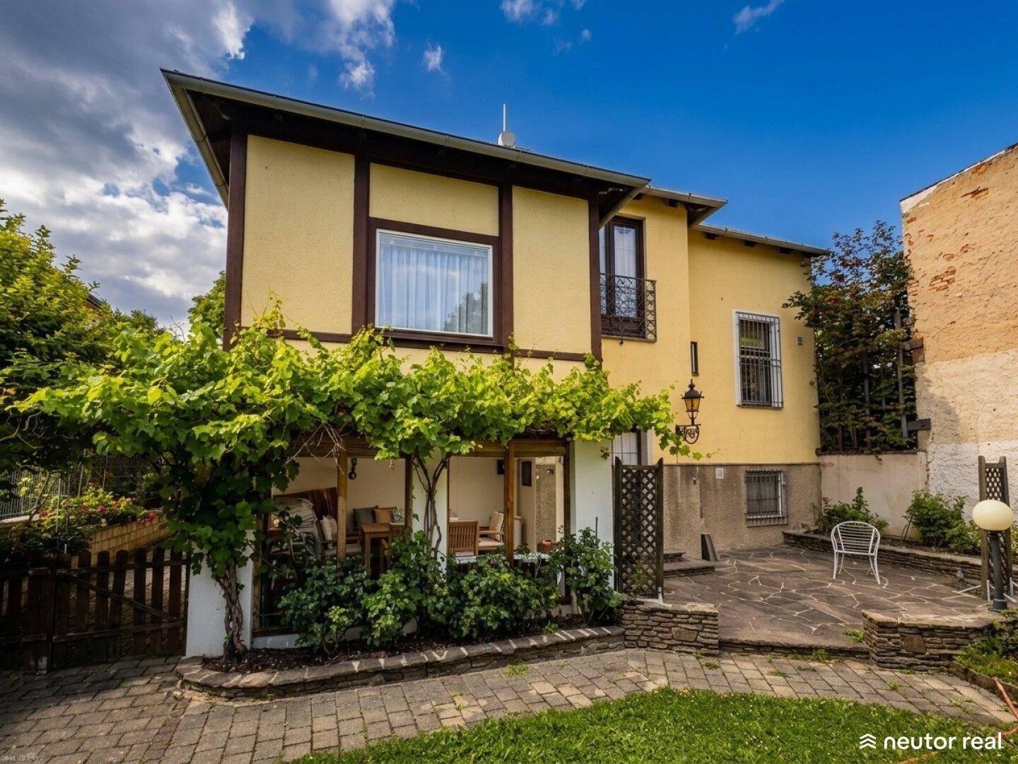 Gelbes Haus mit bewachsener Pergola und gepflastertem Sitzbereich im Garten.