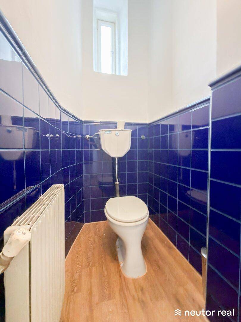 Kleines WC mit blauen Fliesen, Toilette und einem schmalen Fenster für Tageslicht.