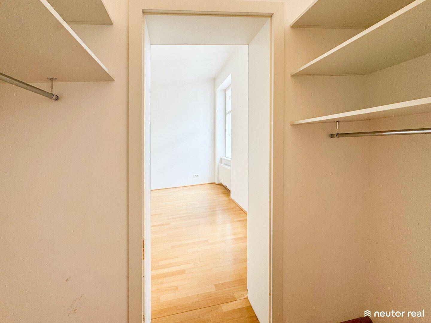 Begehbarer Kleiderschrank mit Regalen und Kleiderstangen, der zu einem hellen Zimmer führt.