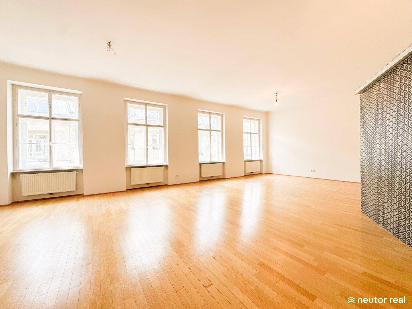 Großes, helles Wohnzimmer mit Holzboden und mehreren hohen Fenstern, die viel Licht hereinlassen.