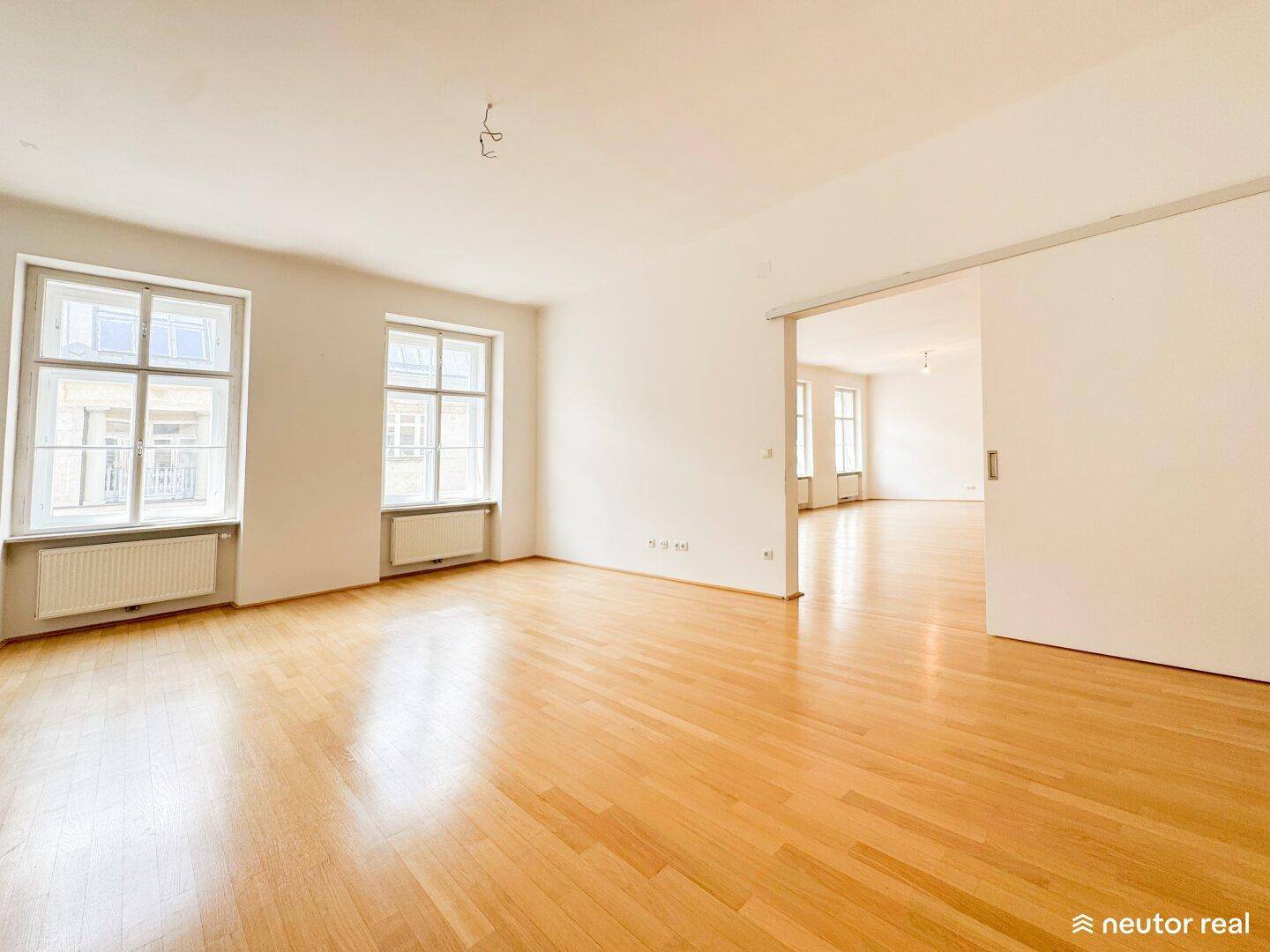Großes, helles Wohnzimmer mit Holzboden und Schiebetür, die zu einem weiteren Raum führt.
