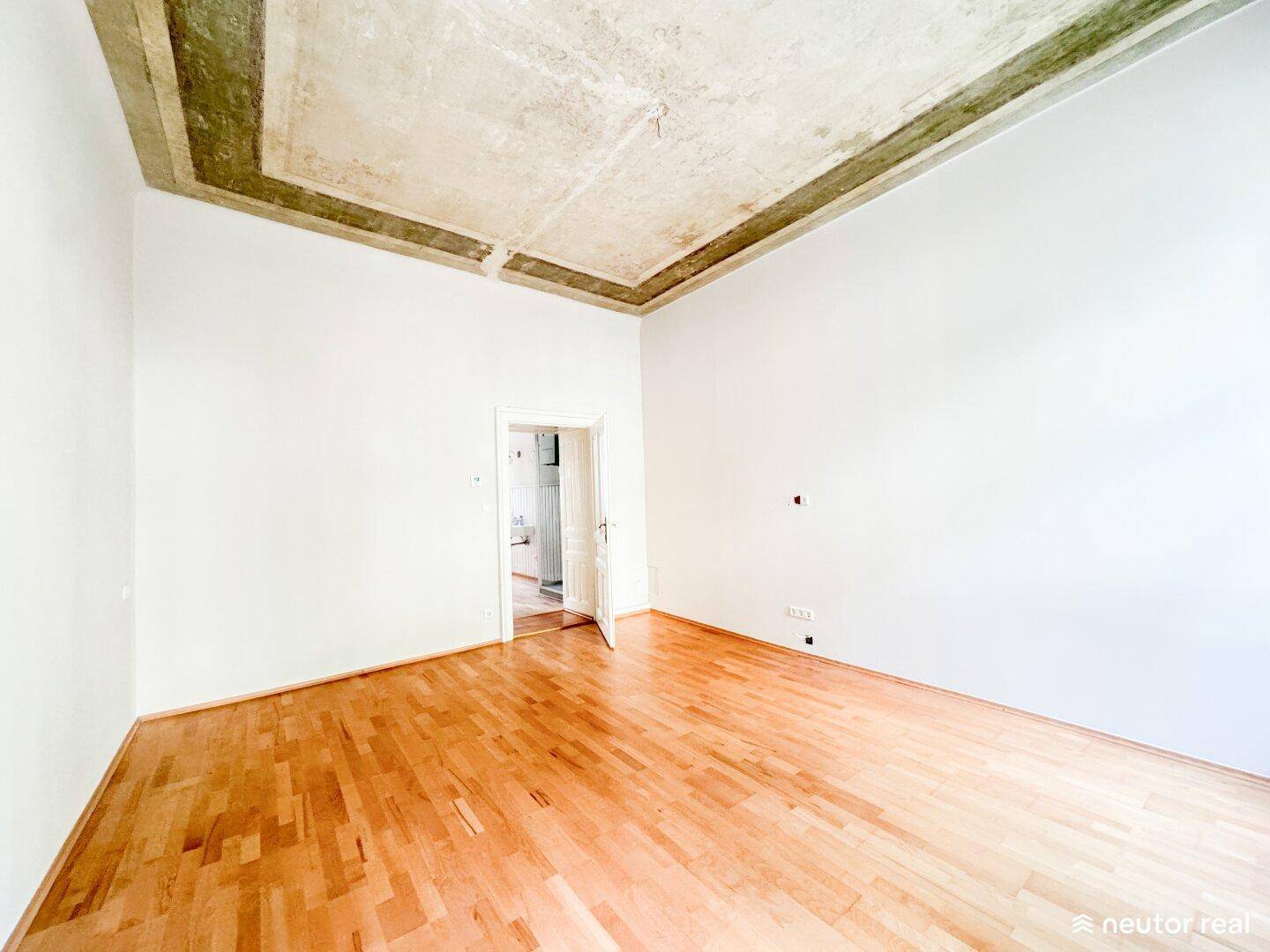 Heller, leerer Raum mit hohen Decken, Holzboden und Stuckelementen an der Decke.