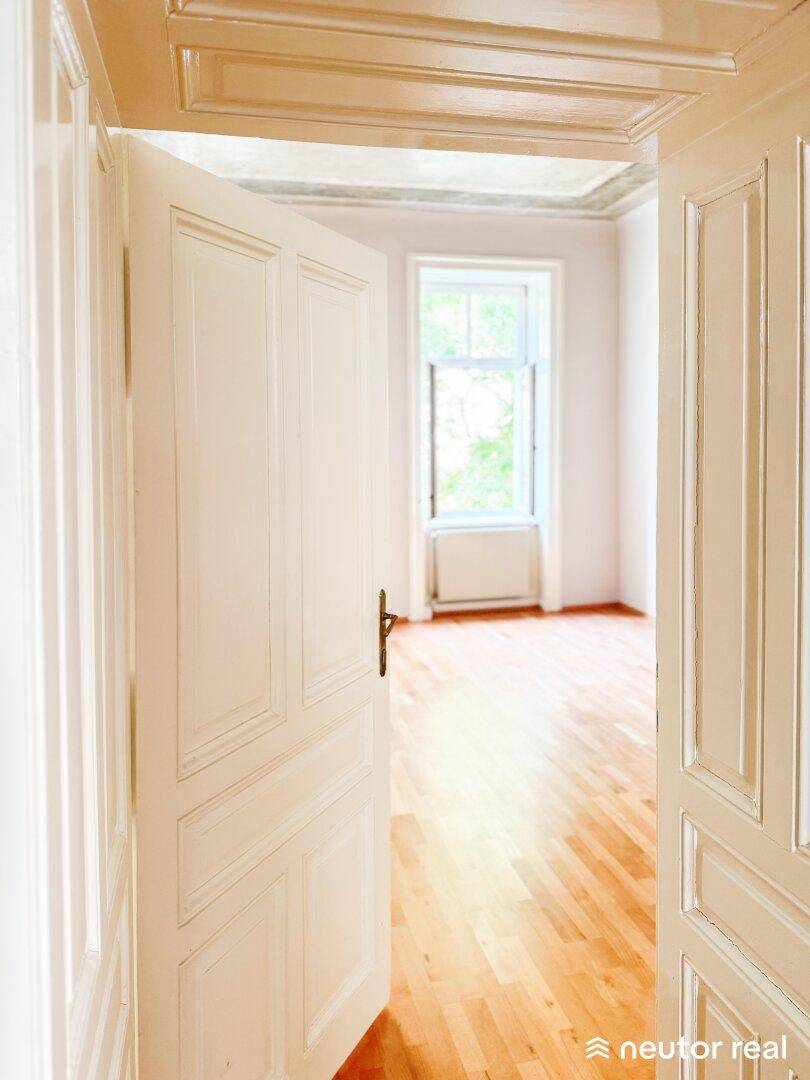 Offene weiße Tür führt in einen weiteren Raum mit Holzboden und großem Fenster.