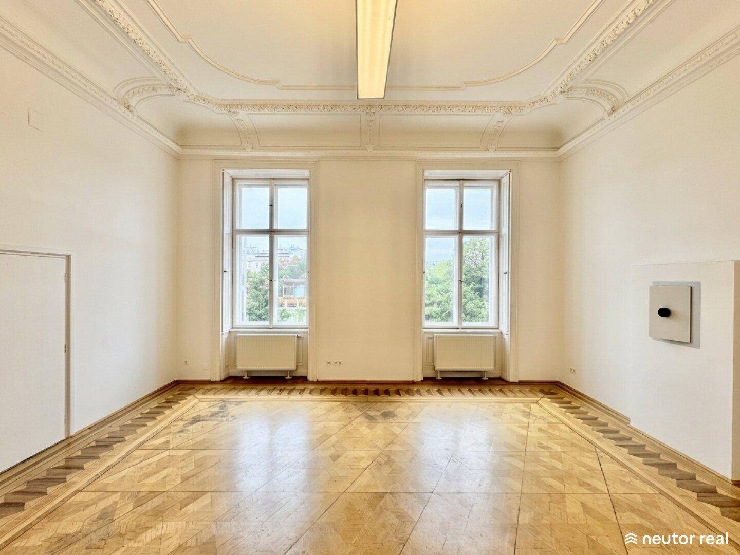 Leerer Büroraum mit Fischgrätparkett, hohen Fenstern und dekorativer Stuckdecke.