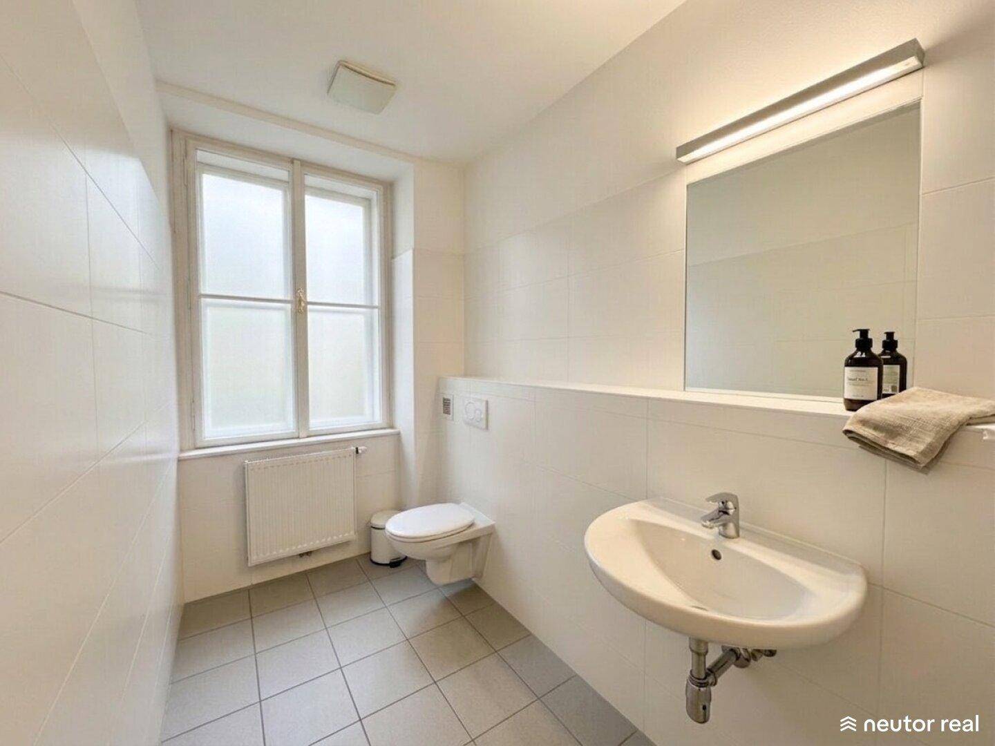 Modernes Badezimmer mit weißen Fliesen, Waschbecken, Toilette und Fenster.