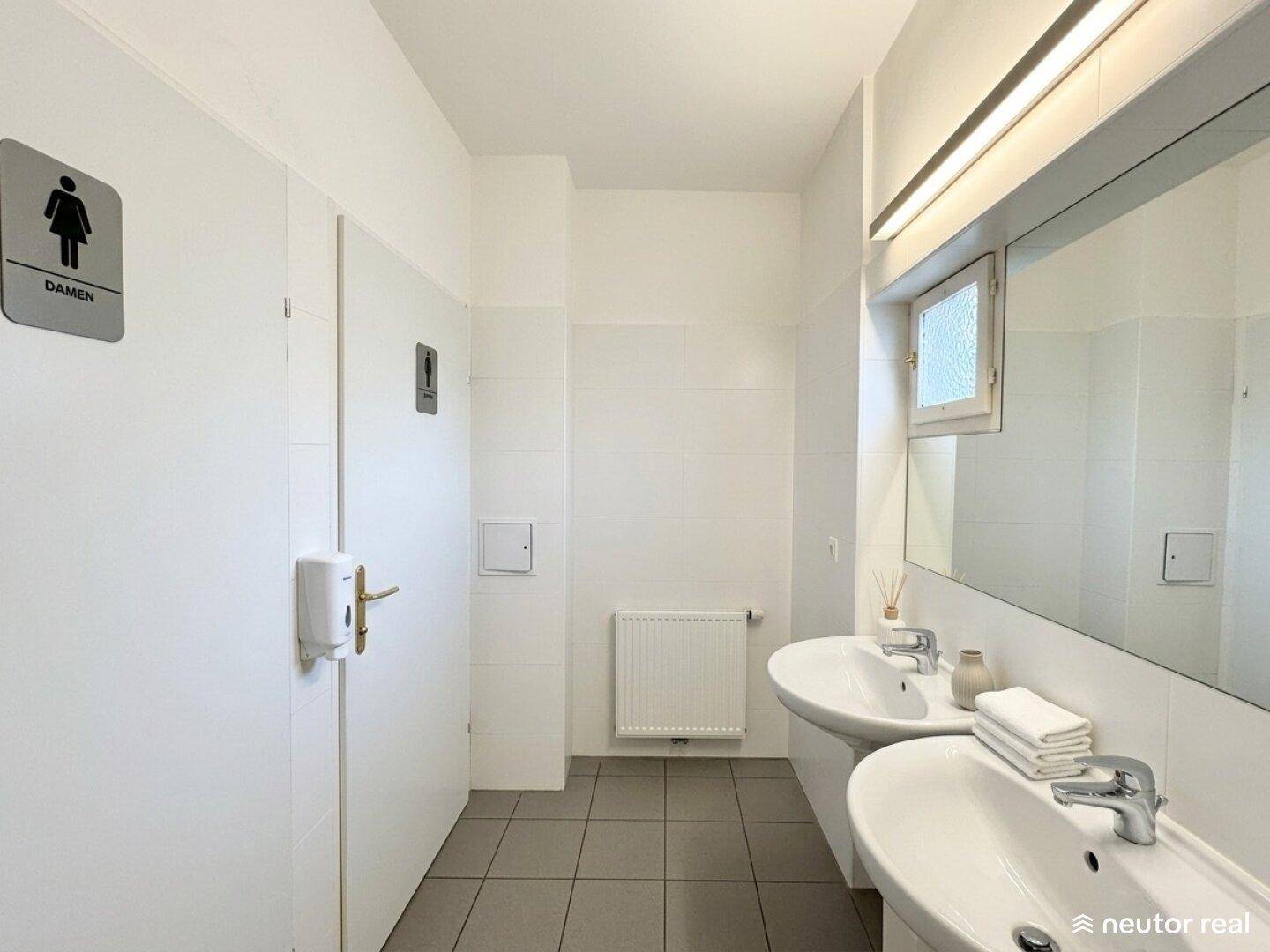 Öffentliche Toilettenanlage mit zwei Waschbecken, Spiegel und einem kleinen Fenster.