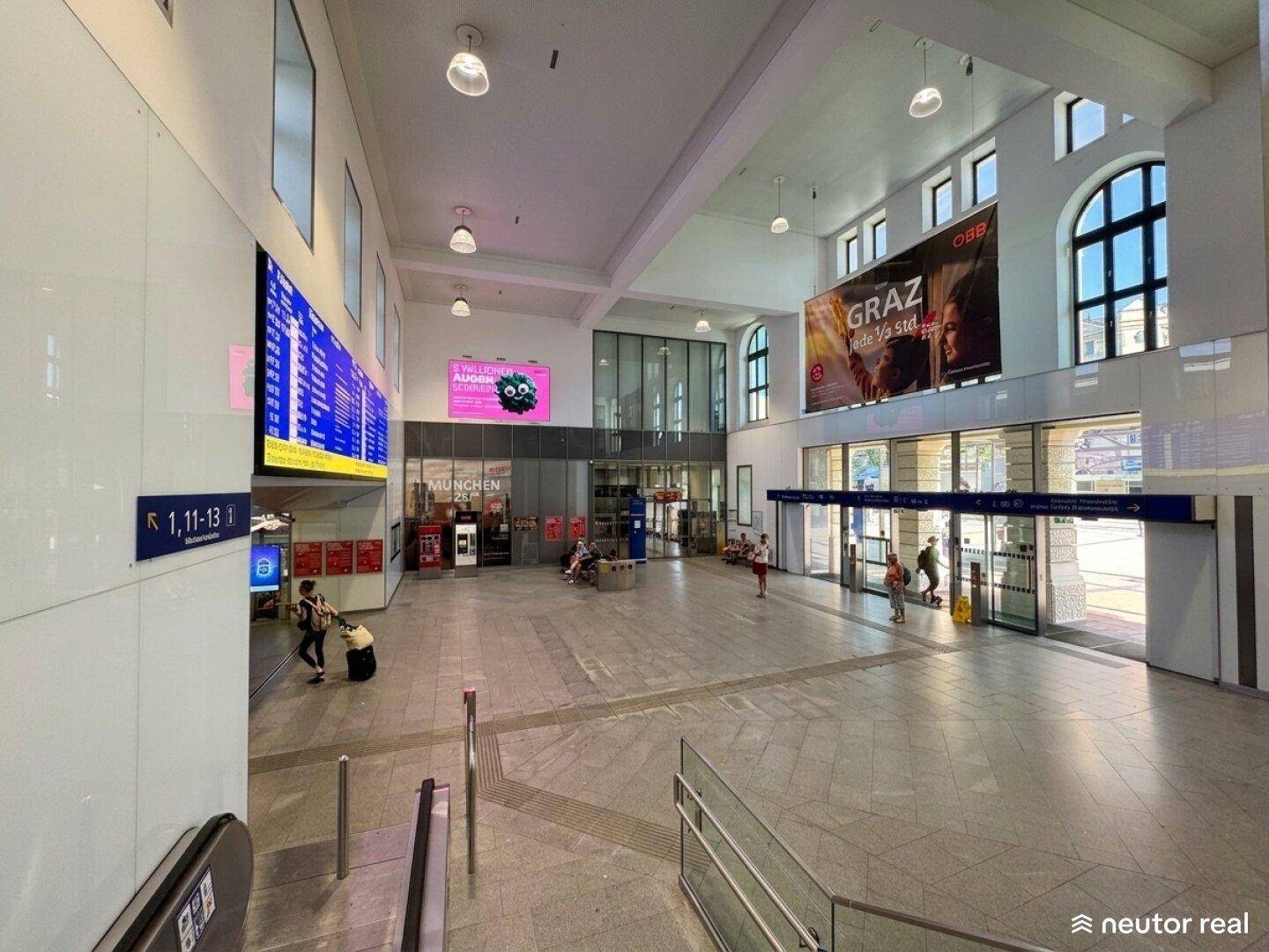Große, helle Bahnhofshalle mit hohen Decken, Anzeigetafeln und vielen Menschen.