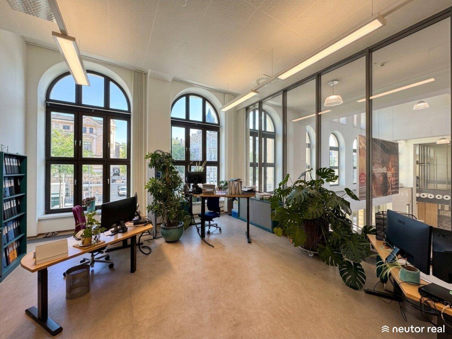 Helles Büro mit großen Fenstern, mehreren Arbeitsplätzen und einer Glaswand zum Flur.