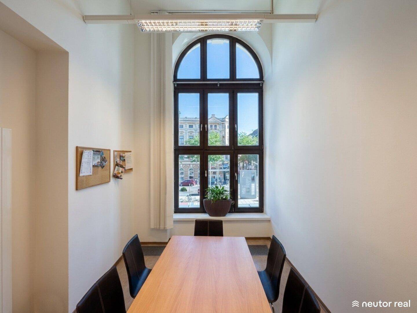 Schmaler Besprechungsraum mit langem Holztisch, Stühlen und einem hohen Rundbogenfenster.