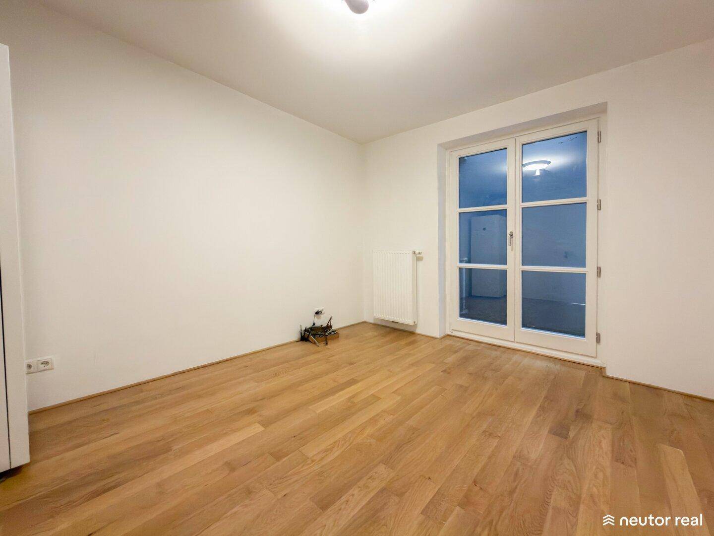 Leeres Zimmer mit hellem Holzparkettboden und großen Fenstertüren, die viel Licht hereinlassen.