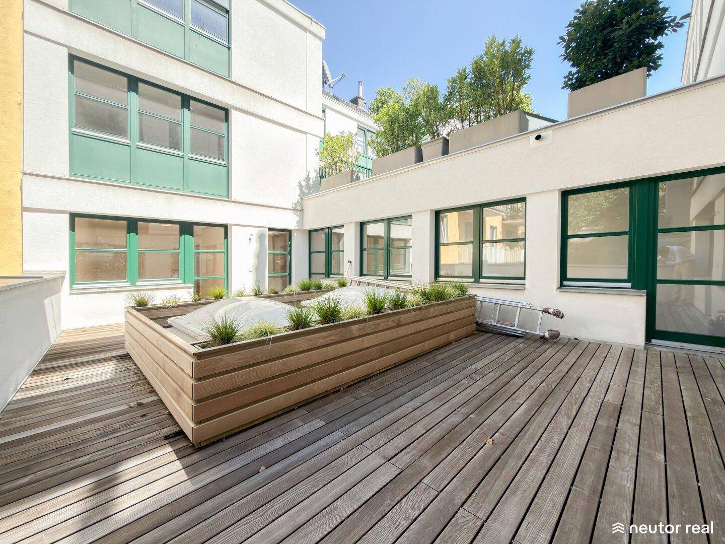 Geräumige Holzterrasse mit Pflanzbeet im Innenhof, umgeben von Gebäudefassaden mit grünen Fenstern.