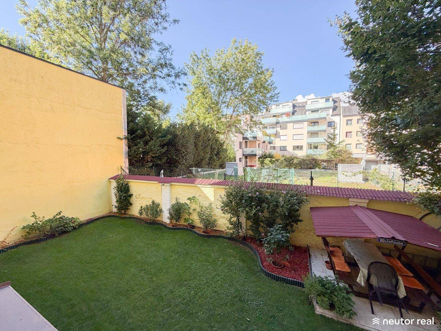 Kleiner, gepflegter Garten mit Rasenfläche und überdachtem Sitzbereich, umgeben von Grün.