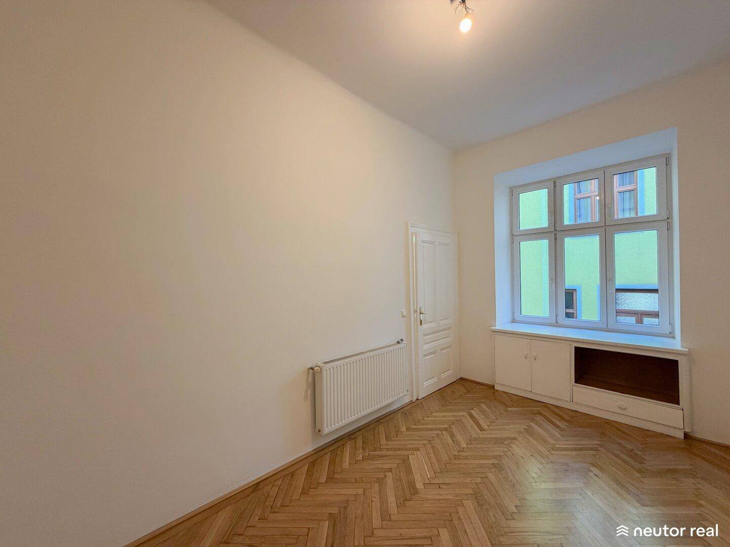 Helles Zimmer mit Fischgrätparkett, Einbauschrank unter dem Fenster und weiß gestrichenen Wänden.