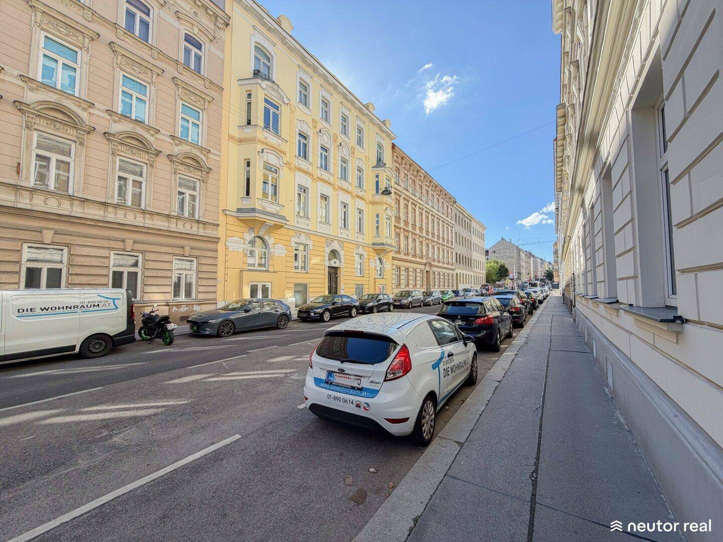 Sonnige Straße mit historischen Gebäuden und geparkten Autos in urbaner Umgebung.
