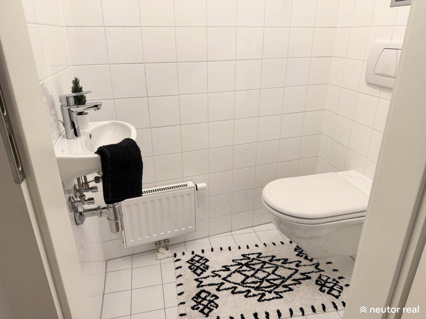 Kleines Gäste-WC mit weißen Fliesen, Wand-WC, Handwaschbecken und Heizkörper.