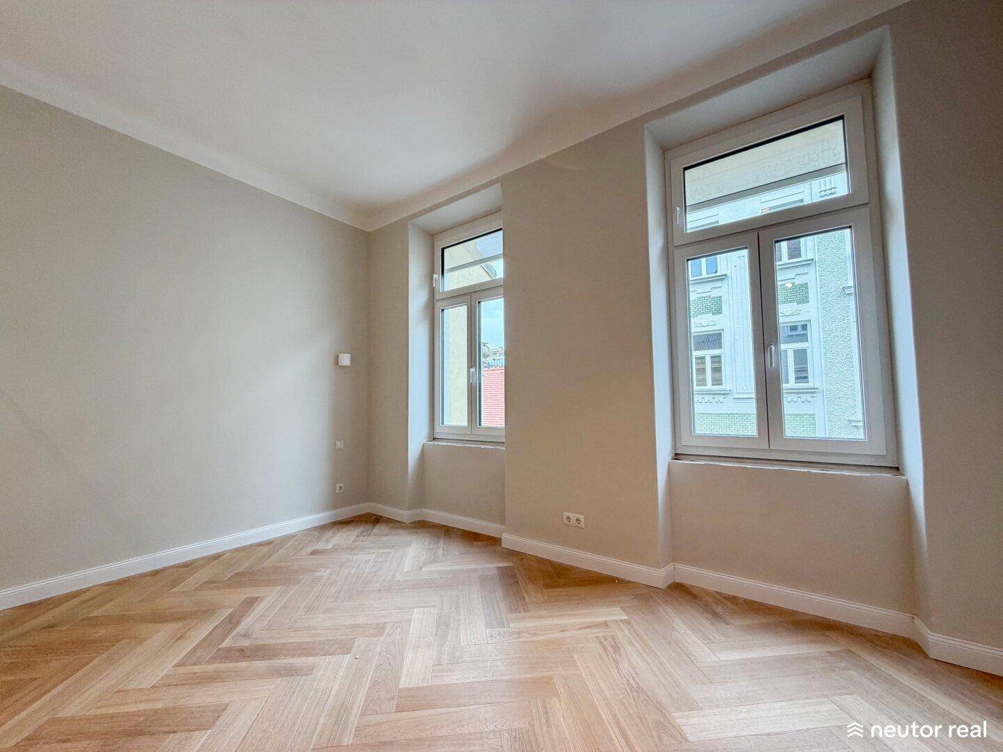 Helles Zimmer mit Fischgrätparkett und großen Fenstern, die viel Tageslicht hereinlassen.