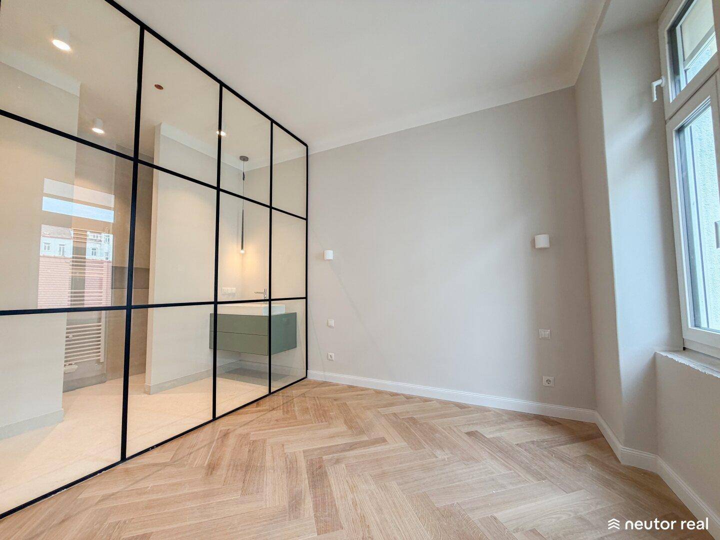 Heller Raum mit Fischgrätparkett und stilvoller Glasabtrennung zum angrenzenden Badezimmer.
