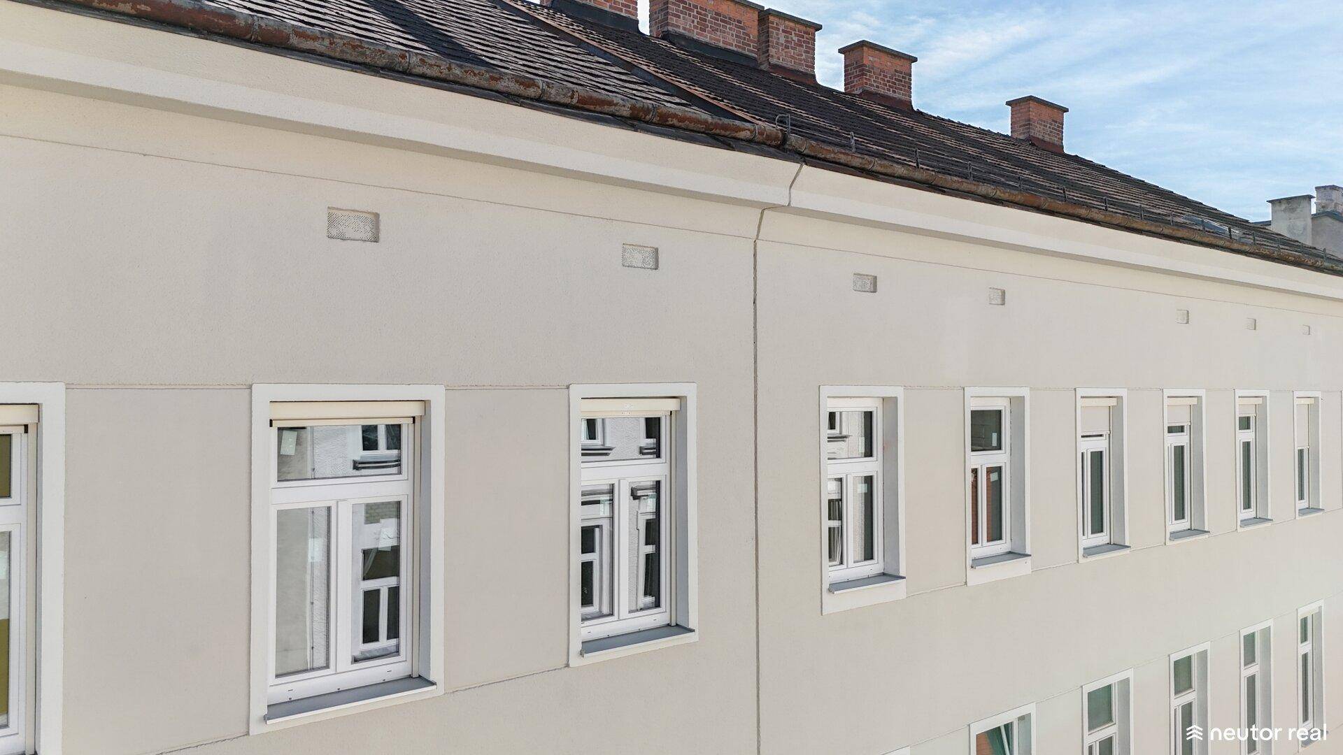 Fassade eines Wohngebäudes mit vielen Fenstern und traditionellem Ziegeldach unter blauem Himmel.